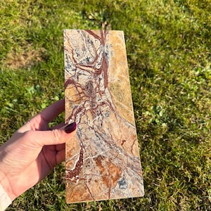 May include: A rectangular stone slab with a natural, earthy pattern. The stone features a mix of tan, brown, red, and gray hues, with intricate veining and patterns. The stone is held against a backdrop of green grass.