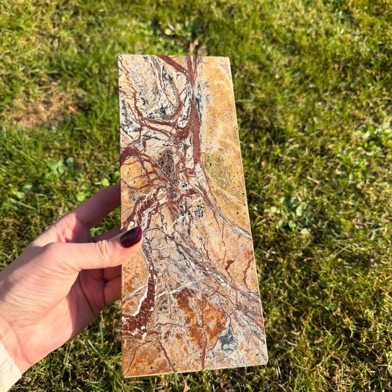 May include: A rectangular stone slab with a natural, earthy pattern. The stone features a mix of tan, brown, red, and gray hues, with intricate veining and patterns. The stone is held against a backdrop of green grass.