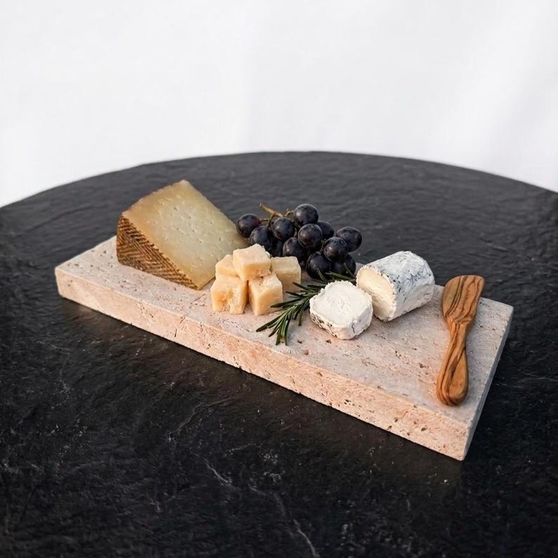May include: A rectangular beige travertine stone serving board holding a wedge of aged cheese, several cubed cheese pieces, a small log of goat cheese, a cluster of dark purple grapes, a sprig of fresh green rosemary, and a wooden cheese spreader.