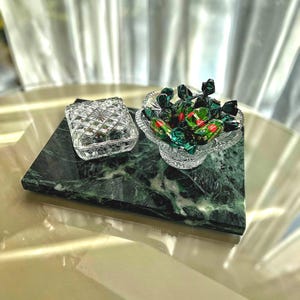 Könnte beinhalten: Eine rechteckige Platte aus dunkelgrünem Marmor, auf der sich eine quadratische, facettierte Glasschatulle mit Deckel sowie eine runde, gewellte Glasschale befinden, die mit mehreren in grün-rote Folie eingewickelten Bonbons gefüllt ist.