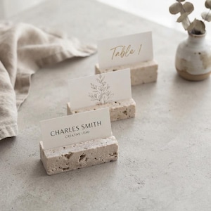 May include: Three rectangular beige stone card holders with rough, porous textures. Each holder supports a white rectangular card. The front card displays the text CHARLES SMITH CREATIVE LEAD, the middle card features a line drawing of a floral sprig, and the back card shows the text Table 1 in gold script.