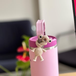 Puede incluir: Un termo rosa claro con un asa y tapa rosas a juego. Una pequeña figura de extraterrestre de ganchillo en beige y blanco está unida a la tapa. El termo es cilíndrico y parece estar hecho de acero inoxidable.