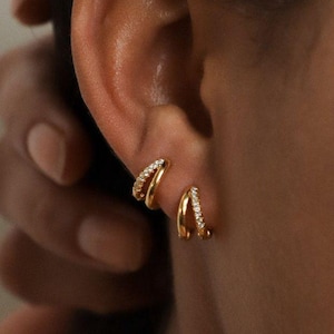 Könnte beinhalten: Goldfarbene Creolen mit kleinen, funkelnden Steinen. Ein Ohrring hat ein Doppelreifen-Design, der andere einen einzelnen Reifen. Die Ohrringe werden an einem Ohr getragen. Schlichtes, elegantes Design, geeignet für den Alltag.