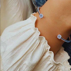 May include: A silver cuff bracelet with two round, clear gemstones. The bracelet is worn on a wrist, and the gemstones are positioned at the ends of the open cuff. The background includes a cream-colored blouse.