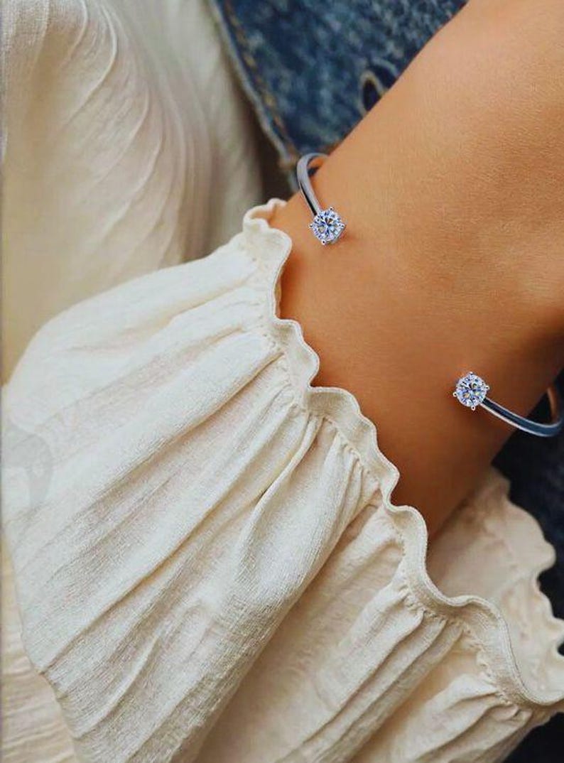 May include: A silver cuff bracelet with two round, clear gemstones. The bracelet is worn on a wrist, and the gemstones are positioned at the ends of the open cuff. The background includes a cream-colored blouse.