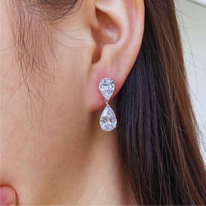 May include: A close-up of a person's ear wearing a pair of sparkling, pear-shaped drop earrings. The earrings are made of clear, faceted stones, likely diamonds or cubic zirconia, and are set in a silver or white metal. The earrings are dangling from the ear.