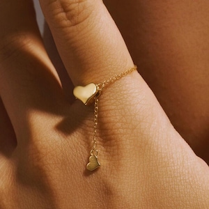 Puede incluir: Un anillo de oro en forma de corazón con un dije de corazón colgante en una cadena delicada.
