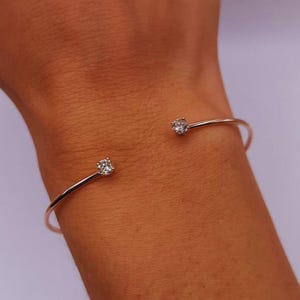 May include: A rose gold cuff bracelet with a minimalist design. The open-ended bracelet features a sparkling, round-cut diamond at each end. The bracelet is shown on a wrist.