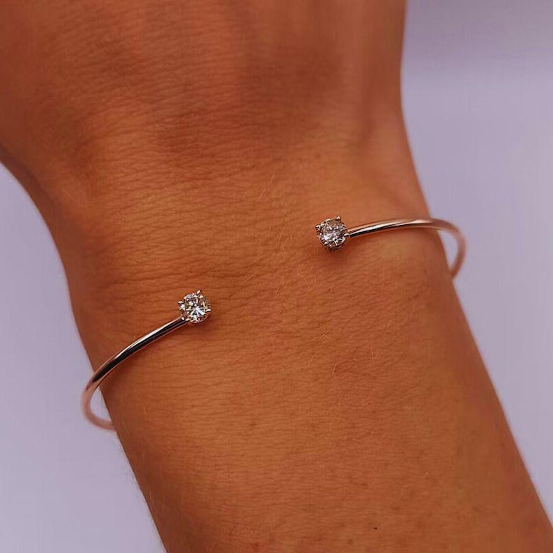 May include: A rose gold cuff bracelet with a minimalist design. The open-ended bracelet features a sparkling, round-cut diamond at each end. The bracelet is shown on a wrist.