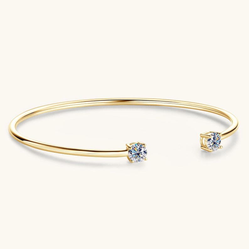 May include: A gold-colored open cuff bracelet with two round, clear gemstones. The bracelet has a simple, elegant design, with the gemstones set at each end of the curved band. The background is a solid, light color.