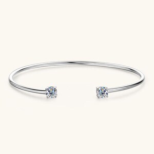 May include: A silver-colored open cuff bracelet with two clear, round-cut gemstones. The bracelet has a simple, elegant design, with the gemstones set at each end of the open band. The gemstones sparkle against the light background.