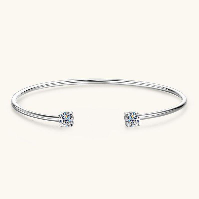May include: A silver-colored open cuff bracelet with two clear, round-cut gemstones. The bracelet has a simple, elegant design, with the gemstones set at each end of the open band. The gemstones sparkle against the light background.