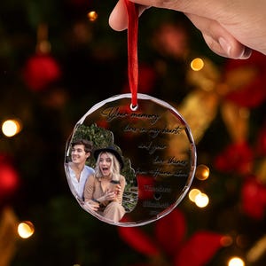 May include: Clear, round Christmas ornament with a photo of a couple and the inscription "Your memory lives in my heart..." held by a red ribbon. The ornament has a textured, faceted edge.