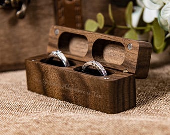 May include: A rectangular dark brown wooden ring box with a hinged lid, featuring two circular slots holding silver rings. The front of the box is engraved with the text My Greatest Adventure is You in a cursive font, showcasing a natural wood grain.