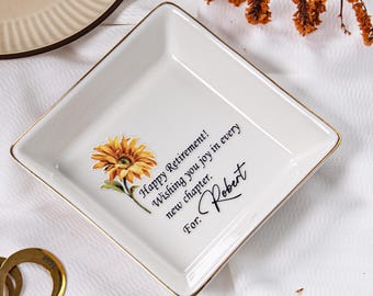 May include: White ceramic square trinket dish with gold trim, featuring a sunflower graphic and the text "Happy Retirement! Wishing you joy in every new chapter. For: Robert."