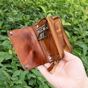 Op de afbeelding: Een bruine leren sleutelhouder met ritssluiting en meerdere sleutelringen. De open sleuteletui toont meerdere sleutels en een kleine leren kwast. De sleutelhouder wordt voor een groene, bladerrijke achtergrond gehouden.