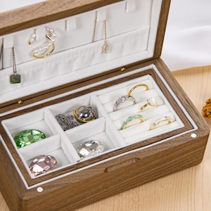 May include: Open wooden jewellery box with a light-coloured interior and various rings, necklaces, and earrings. The box is made of dark wood and has a hinged lid. The jewellery includes gold, silver, and gemstone pieces.