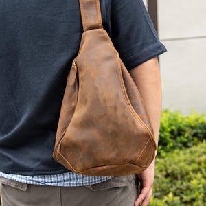 May include: A brown leather sling bag with a zipper closure and adjustable strap. The bag is worn over the shoulder and across the chest. The leather has a distressed look.