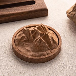 May include: A round, wooden coaster with a detailed mountain range carving. The coaster is made of a light brown wood and features a raised relief of peaks and valleys. The coaster is approximately 10 cm in diameter.