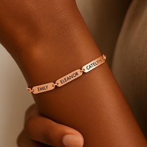 Könnte beinhalten: Ein roségoldenes Armband mit drei rechteckigen Namensschildern. Die Namen "EMILY", "ELEANOR" und "CATELYN" sind in schwarzer Schrift eingraviert. Das Armband wird am Handgelenk getragen.