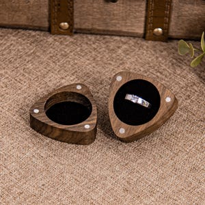 May include: A silver ring with a square-cut central stone sits in an open, triangular wooden ring box with a black velvet interior. An empty box of the same design is next to it. The boxes have inlaid white accents. The ring box is made of wood.