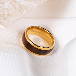 May include: A gold ring with a dark brown wood inlay. The ring is displayed on a white fabric, accentuating its polished appearance. The design features a band of dark wood encased in a gold metal frame, creating a striking contrast.