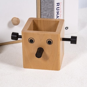 Op de afbeelding: Een lichtgekleurde houten bureau-organizer in de vorm van een gezicht, met zwarte accenten voor ogen, een neus en handvatten. De organizer is een kubus met een holle binnenkant voor het opbergen van pennen, potloden of andere kleine items. Het ontwerp is grillig en speels.