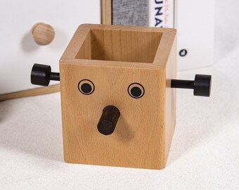 May include: A light-colored wooden desk organizer shaped like a face, with black accents for eyes, a nose, and handles. The organizer is a cube with a hollow interior for storing pens, pencils, or other small items. The design is whimsical and playful.