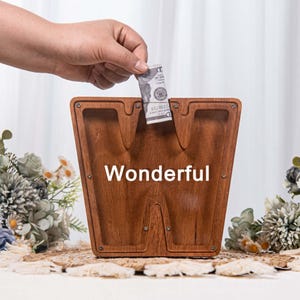 May include: A wooden money bank shaped like the letter "W" with the word "Wonderful" printed on it. A hand is inserting a dollar bill into the slot. The bank has a warm brown color and is decorated with flowers.