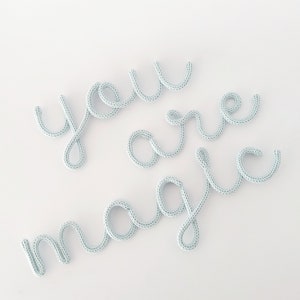 May include: Light blue knitted text on a white background spells out "you are magic" in a cursive font. The letters are made of a soft, textured material, creating a whimsical and decorative design.