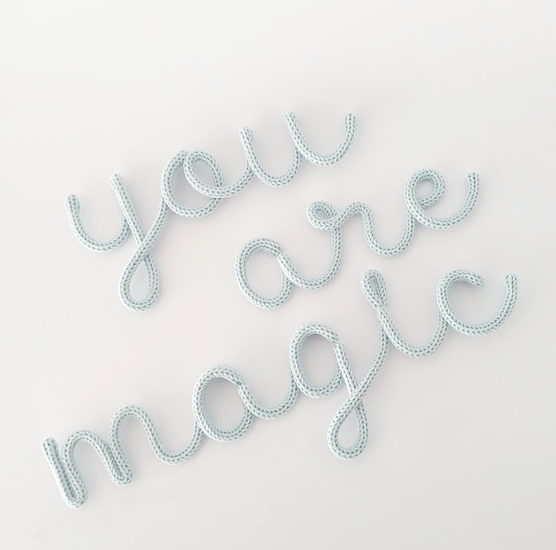 May include: Light blue knitted text on a white background spells out "you are magic" in a cursive font. The letters are made of a soft, textured material, creating a whimsical and decorative design.