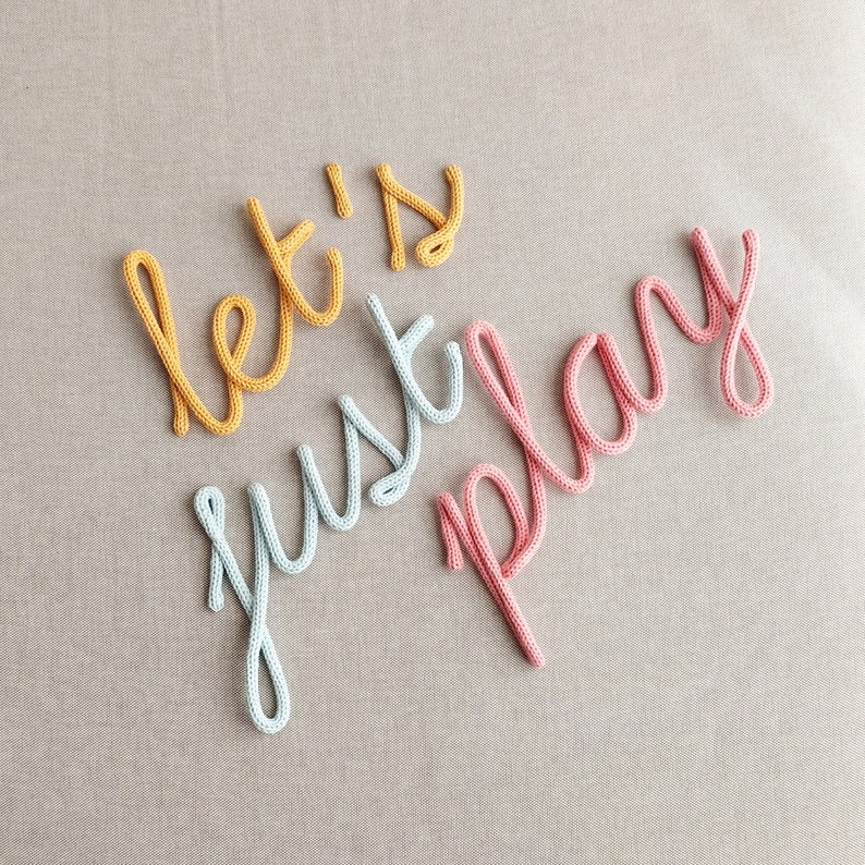 K&ouml;nnte beinhalten: Nahaufnahme der Worte "let's just play" aus gestricktem Garn. Die W&ouml;rter sind in Schreibschrift gehalten, mit "let's" in Gelb, "just" in Hellblau und "play" in Rosa. Der Hintergrund ist ein neutraler, hellbrauner Stoff.