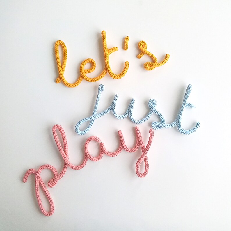 K&ouml;nnte beinhalten: Auf wei&szlig;em Hintergrund steht der Schriftzug "let's just play" aus gestricktem Garn. Das Wort "let's" ist gelb, "just" ist hellblau und "play" ist rosa. Das Garn ist dick und die Buchstaben sind in Schreibschrift gehalten.