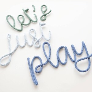 K&ouml;nnte beinhalten: Wanddekoration mit dem Schriftzug "let's just play" in einer verspielten Schriftart. Die W&ouml;rter sind aus dickem, gestricktem Material in Gr&uuml;n, Hellblau und Blau gefertigt, vor einem wei&szlig;en Hintergrund. Ein charmantes Element f&uuml;r ein Kinderzimmer oder Spielzimmer.