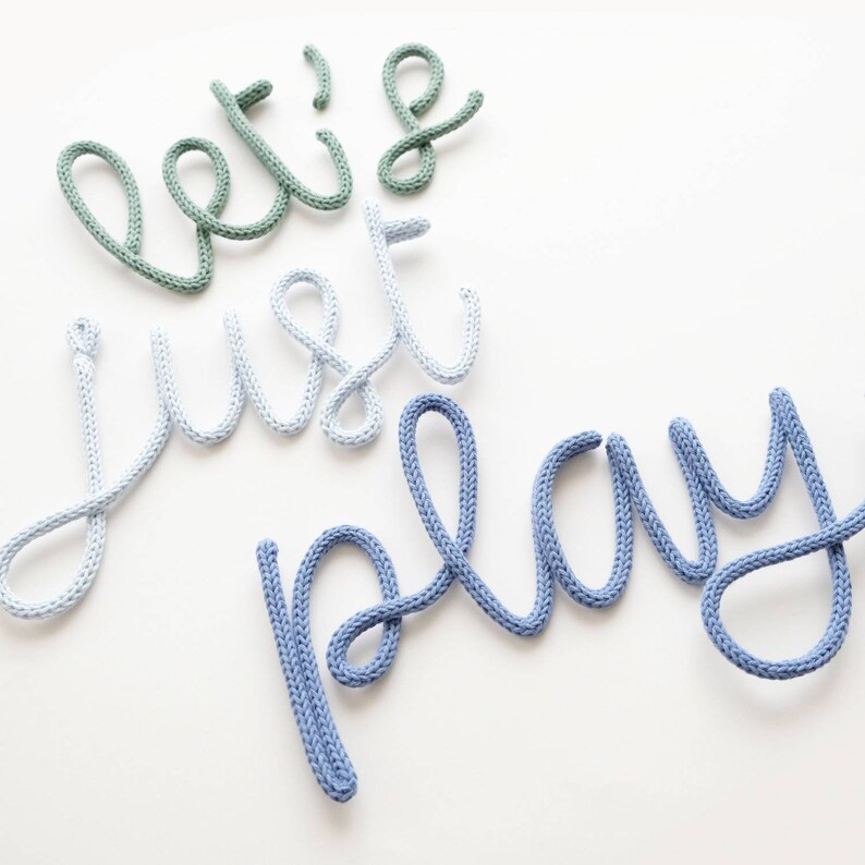 K&ouml;nnte beinhalten: Wanddekoration mit dem Schriftzug "let's just play" in einer verspielten Schriftart. Die W&ouml;rter sind aus dickem, gestricktem Material in Gr&uuml;n, Hellblau und Blau gefertigt, vor einem wei&szlig;en Hintergrund. Ein charmantes Element f&uuml;r ein Kinderzimmer oder Spielzimmer.