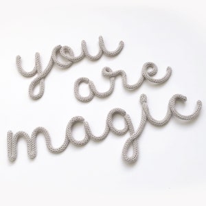 May include: A light gray knitted wall art piece spelling out "you are magic" on a white background. The letters are in a cursive style, with a soft, textured appearance. The overall design is simple and elegant.