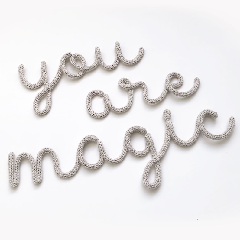 May include: A light gray knitted wall art piece spelling out "you are magic" on a white background. The letters are in a cursive style, with a soft, textured appearance. The overall design is simple and elegant.