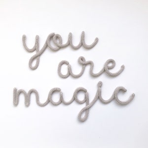 May include: A light gray knitted wall hanging spelling out the words "you are magic" in a cursive font. The letters are made of a thick, textured yarn and are displayed against a white background.