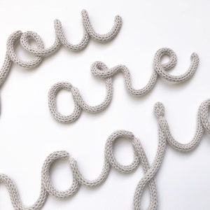 May include: Close-up of the word "joy" spelled out in gray knitted yarn on a white background. The letters are in a cursive style, with a soft, textured appearance. The word is arranged in a playful, artistic manner.