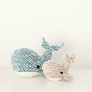 May include: Two plush whale toys. One is light blue with a white belly, the other is beige with a white belly. Both have black eyes and are made of a soft, textured material. The larger whale has a tail fin.