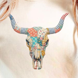 Puede incluir: Camiseta color crema con un diseño de calavera de toro en patchwork. La calavera está compuesta por varios patrones de tela, incluyendo florales, cuadros y lunares, con acentos azules, rojos y amarillos. Los cuernos son a cuadros azules y rojos.