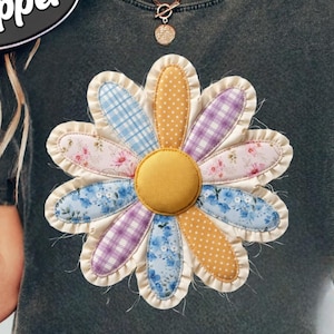 Puede incluir: Camiseta gris oscuro con una gran margarita aplicada. Los pétalos de la margarita son de varias telas estampadas: floral azul, lunares amarillos y cuadros morados. La camiseta también tiene el texto "COMFORT COLORS Pepper" en un óvalo blanco.