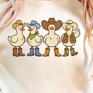 May include: A cream-coloured t-shirt with a cartoon illustration of four ducks dressed as cowboys. Each duck wears a different coloured cowboy hat, boots, and accessories, including a vest and bandana. The ducks are standing side by side.
