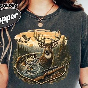 Puede incluir: Camiseta gris oscuro con un diseño de temática natural. El diseño incluye un ciervo, un pez, un rifle y un paisaje montañoso con pájaros. El texto "COMFORT COLORS Pepper" está impreso en la parte superior izquierda.