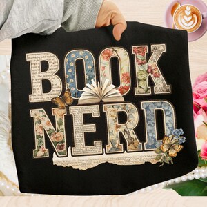 Può includere: T-shirt nero con la scritta "BOOK NERD" in un collage di disegni floreali e fantasia. Le lettere sono decorate con fiori, farfalle e pagine di libri. Un libro aperto è al centro.