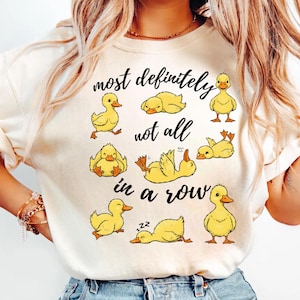 Puede incluir: Camiseta color crema con un diseño lúdico de patitos amarillos en varias poses. El texto "most definitely not all in a row" está impreso en negro. La camiseta está hecha de un material suave.