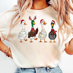 Könnte beinhalten: Cremefarbenes T-Shirt mit einem festlichen Design von vier Enten. Jede Ente ist mit Weihnachtsbeleuchtung und Accessoires geschmückt, darunter Weihnachtsmützen und Rentiergeweihe. Die Enten sind im Cartoon-Stil dargestellt und schaffen eine fröhliche und saisonale Ästhetik.