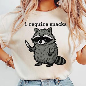 May include: Cream-colored t-shirt featuring a cartoon raccoon holding a knife. The raccoon is gray and black with a white stripe across its eyes. The text "i require snacks" is printed above the raccoon in black.