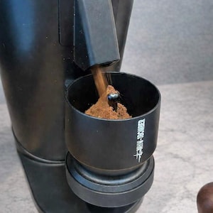 Puede incluir: Un molinillo de café negro con la parte superior plateada. El café molido se vierte en un recipiente negro. El recipiente tiene el texto "FELLOW-SPOUTED" impreso en blanco.