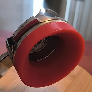 May include: A red and silver tea infuser with a perforated interior. The infuser has a wide, rounded opening and a handle. It is resting on a round, light-colored wooden surface.